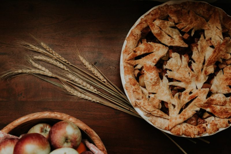 A delicious looking pie, with wheat and apples on the table.